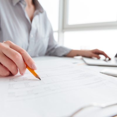 Close up portrait of a woman holding pencil over paper documents while sitting and working with laptop computer at home